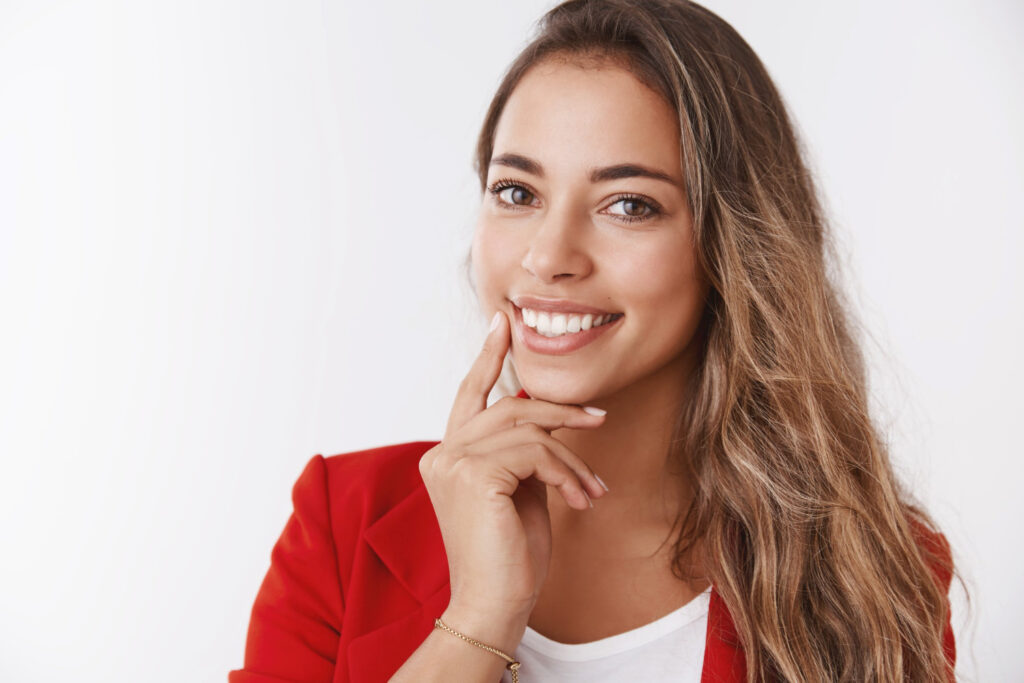 Mulher jovem sorridente de blazer vermelho toca o rosto, exibindo um sorriso saudável após tratamento ATM em São José dos Campos.
